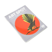 The Bright Fantail Art Spot is a round, repositionable wall art print showing a bird with a fanned tail on a branch, set against a bright red background. It has a matte fabric finish, comes in ART SPOT packaging, and is made in New Zealand.