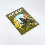 A flat-lay image of the Botanical Tui Art Spot print shows two birds among yellow flowers and greenery in a clear sleeve. This repositionable wall art uses unique adhesive, is biodegradable, compostable, and made in New Zealand.