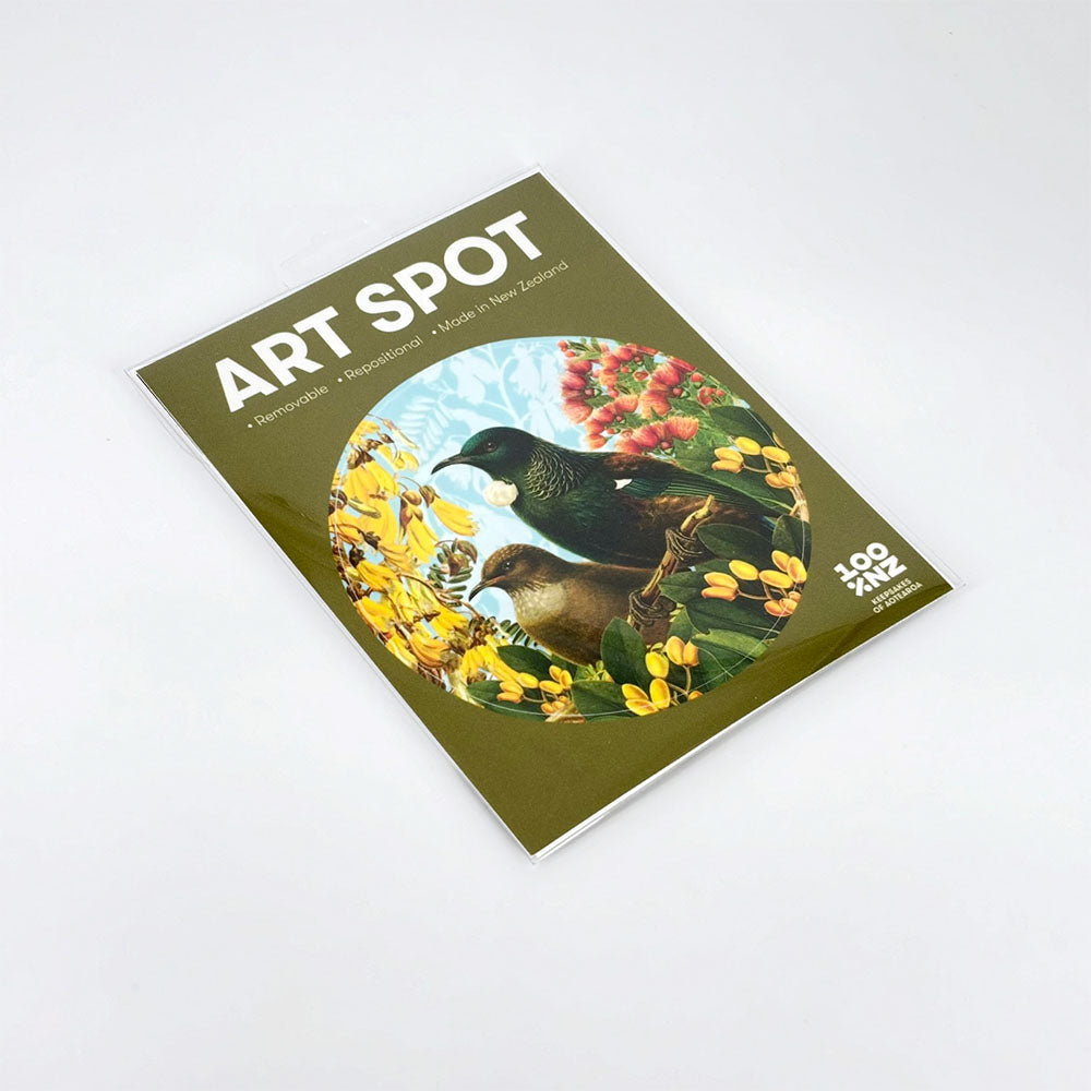 A flat-lay image of the Botanical Tui Art Spot print shows two birds among yellow flowers and greenery in a clear sleeve. This repositionable wall art uses unique adhesive, is biodegradable, compostable, and made in New Zealand.