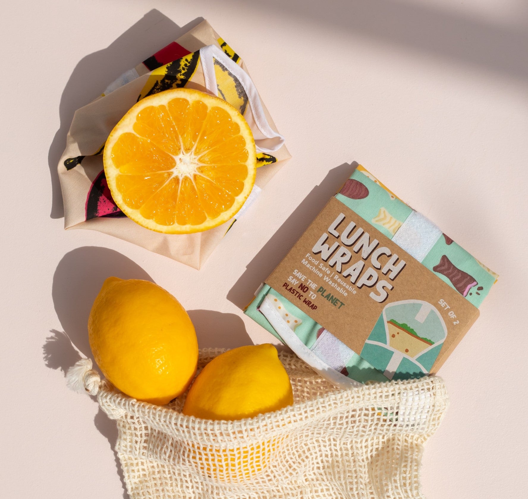 A halved orange on patterned cloth, two whole lemons in a mesh bag, and a package labeled Chocolate Fish Lunch Wrap with reusable, food-safe certified wraps featuring a colorful design are arranged on a light surface.