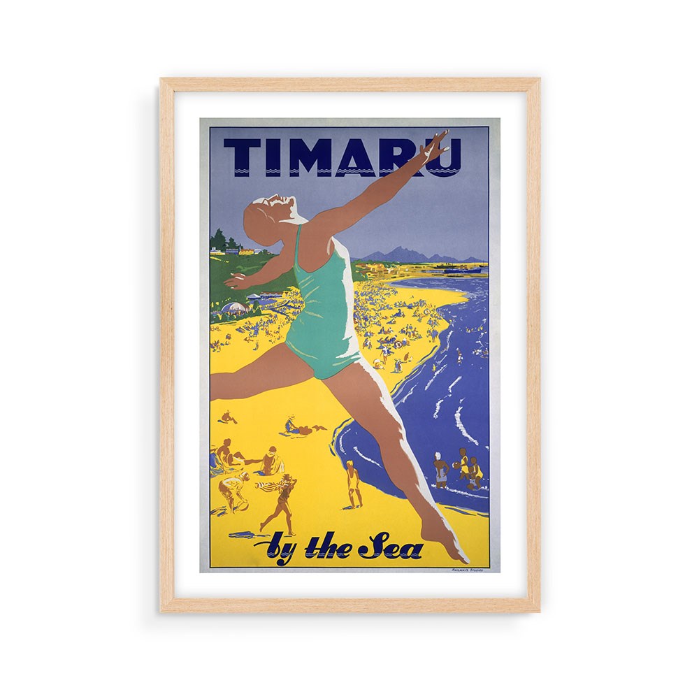 The Timaru Tourist Print is a vintage-style poster in a light wood frame, showing a person in a turquoise swimsuit leaping on a yellow beach with people and mountains behind, inspired by classic tourism posters. Text reads: Timaru by the Sea.