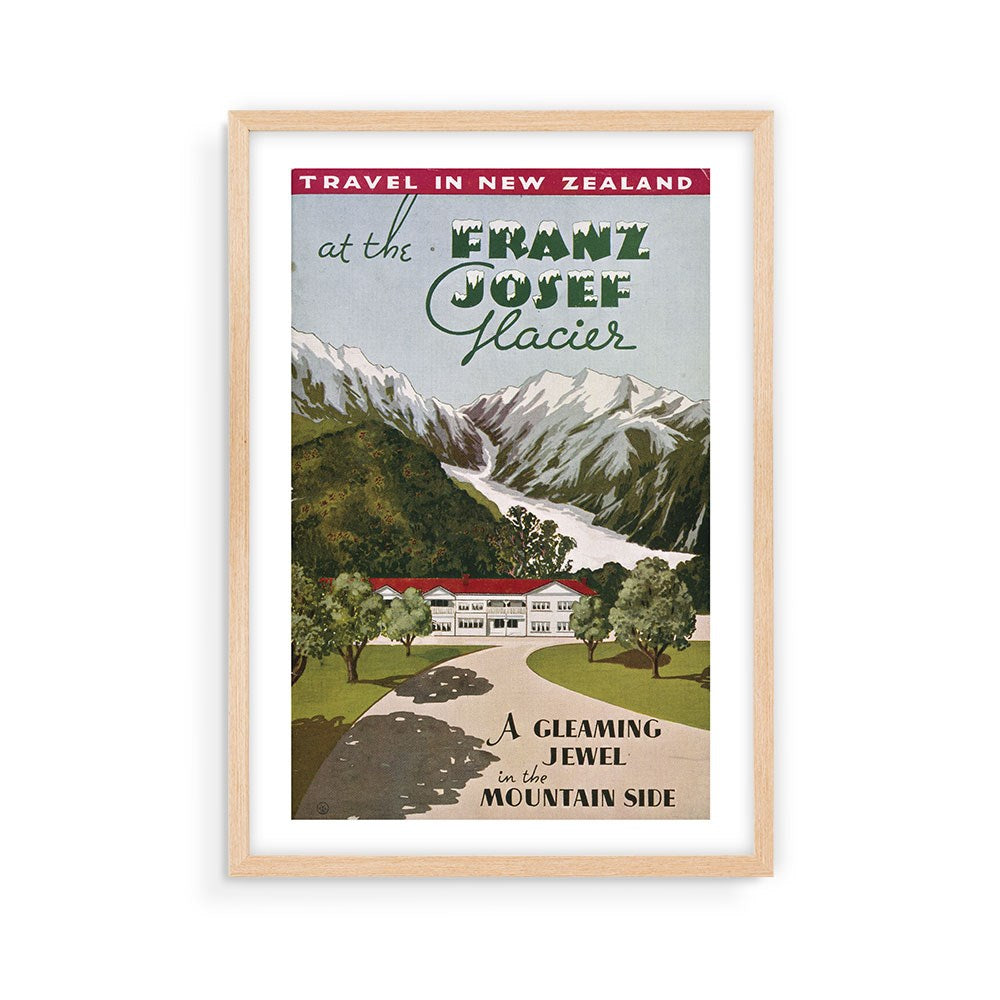 The Franz Josef Glacier Tourist Print is a framed vintage travel poster of New Zealand's Franz Josef Glacier, featuring snowy peaks, a river, trees, and a building with the phrase “A gleaming jewel on the mountain side.”.