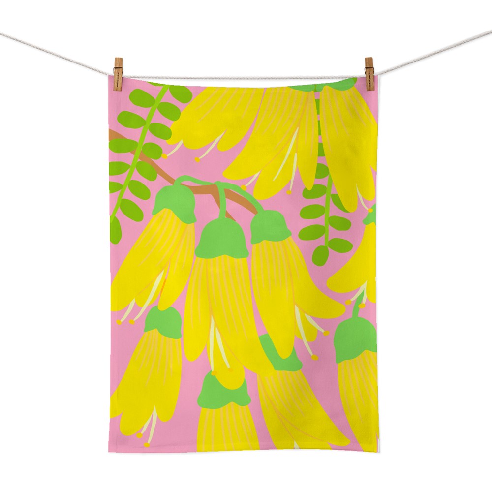 The Bright Botanical Kowhai Tea Towel, screen printed on cotton with yellow kowhai flowers and green leaves on a pink background, hangs from a clothesline with two wooden clothespins.