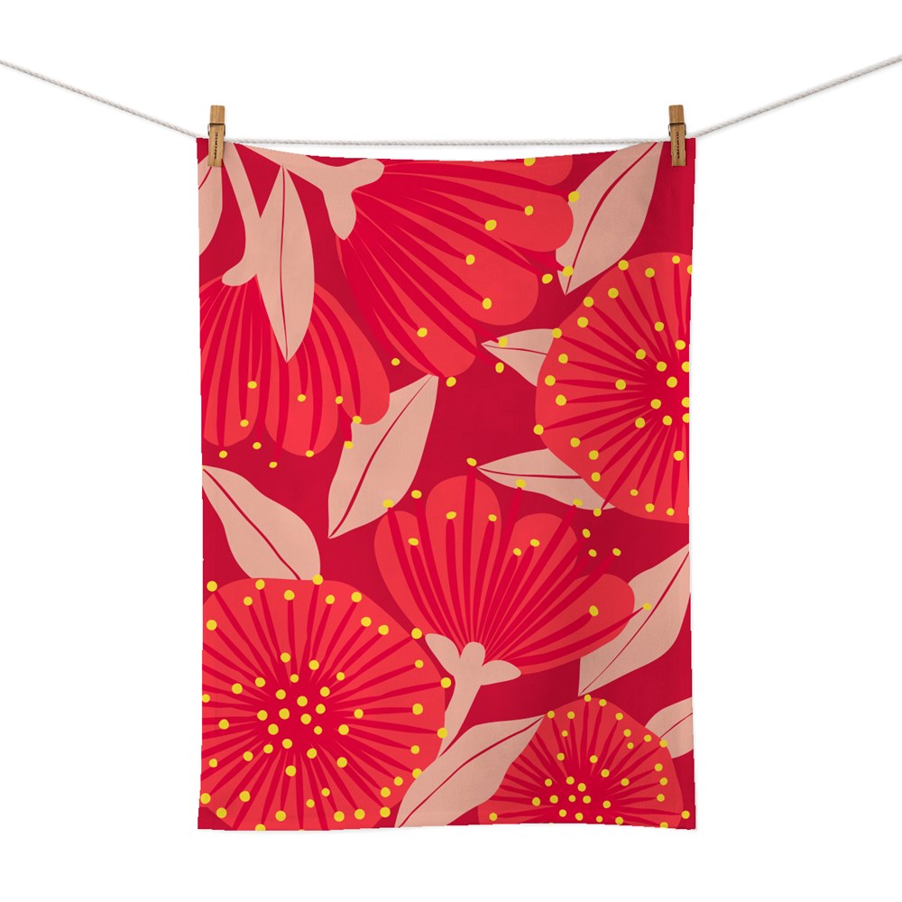 The Bright Botanical Red Pohutukawa Tea Towel is a rectangular cotton tea towel with a red background, decorated with bold red and pink botanical flowers, yellow dots, and pale pink leaves. It hangs on a line with two wooden clothespins.