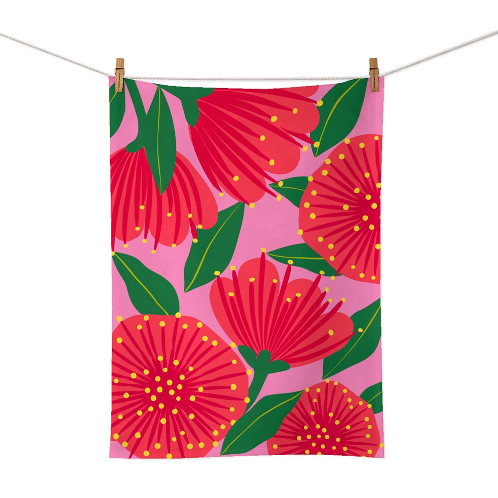 The Bright Botanical Pink Pohutukawa Tea Towel, screen printed on cotton in New Zealand, displays bold red pohutukawa flowers, green leaves, and yellow dots on a pink background. It’s pictured hanging from a clothesline with wooden clothespins.