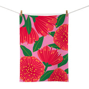 The Bright Botanical Pink Pohutukawa Tea Towel, screen printed on cotton in New Zealand, displays bold red pohutukawa flowers, green leaves, and yellow dots on a pink background. It’s pictured hanging from a clothesline with wooden clothespins.