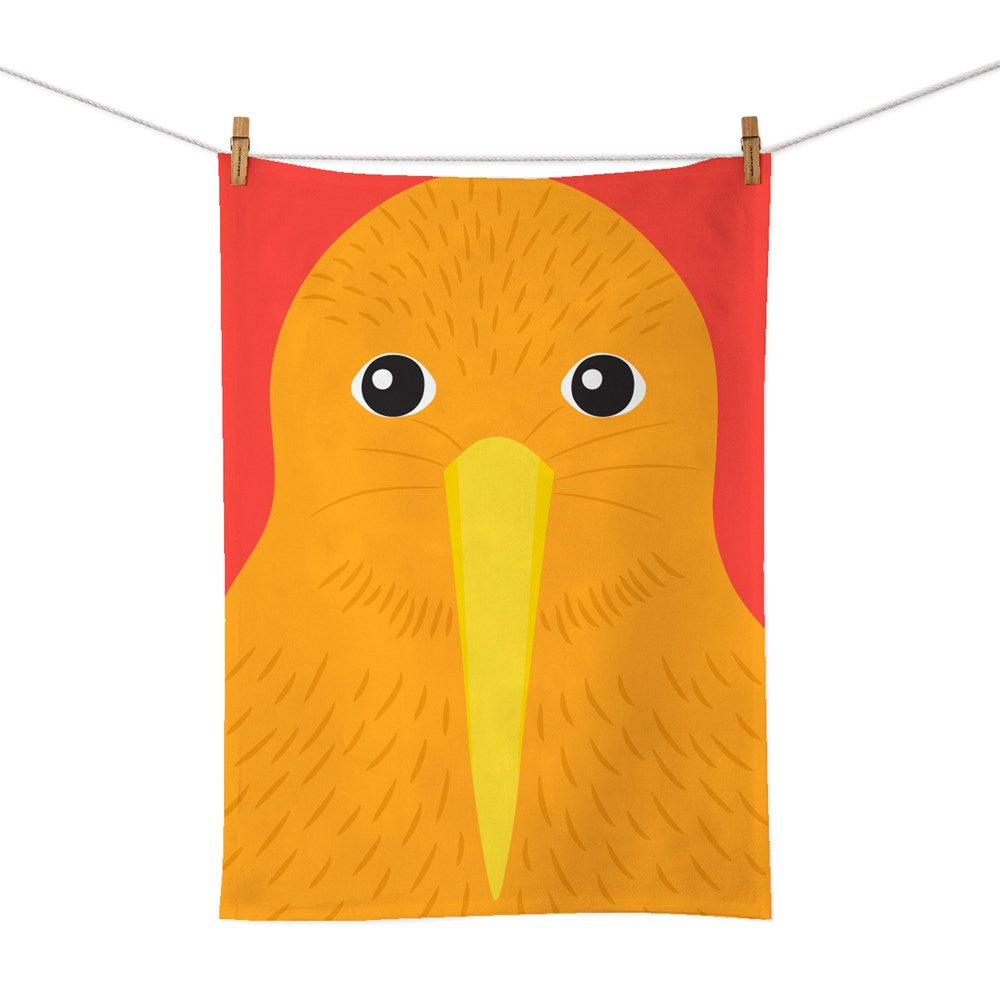 The Bright Kiwi Tea Towel is an orange rectangular cotton towel with a playful cartoon kiwi bird face and beak, hanging by clothespins against a red background, celebrating New Zealand-inspired full colour design.