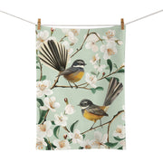 The Chatty Fantails Tea Towel is a cotton tea towel, screen printed and designed in New Zealand, featuring two fantail birds perched on blossoming branches with white flowers and green leaves.