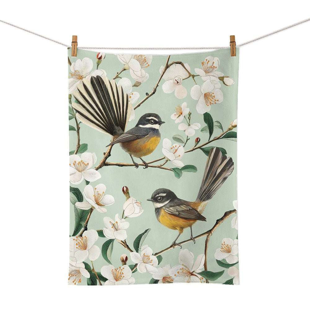 The Chatty Fantails Tea Towel is a cotton tea towel, screen printed and designed in New Zealand, featuring two fantail birds perched on blossoming branches with white flowers and green leaves.
