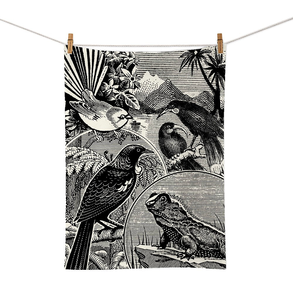 The NZ Stamp Tea Towel features a black and white illustration of four birds on branches and a frog amid foliage and mountains, screen printed on cotton. Designed in New Zealand, its shown clipped to a line with clothespins.