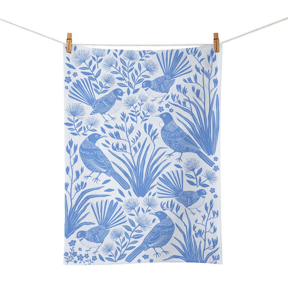 The Birdsong Blues Tea Towel is a screen-printed cotton towel with a blue and white botanical design featuring stylized birds among plants, inspired by New Zealand’s nature, shown hanging on a clothesline with two wooden clothespins.
