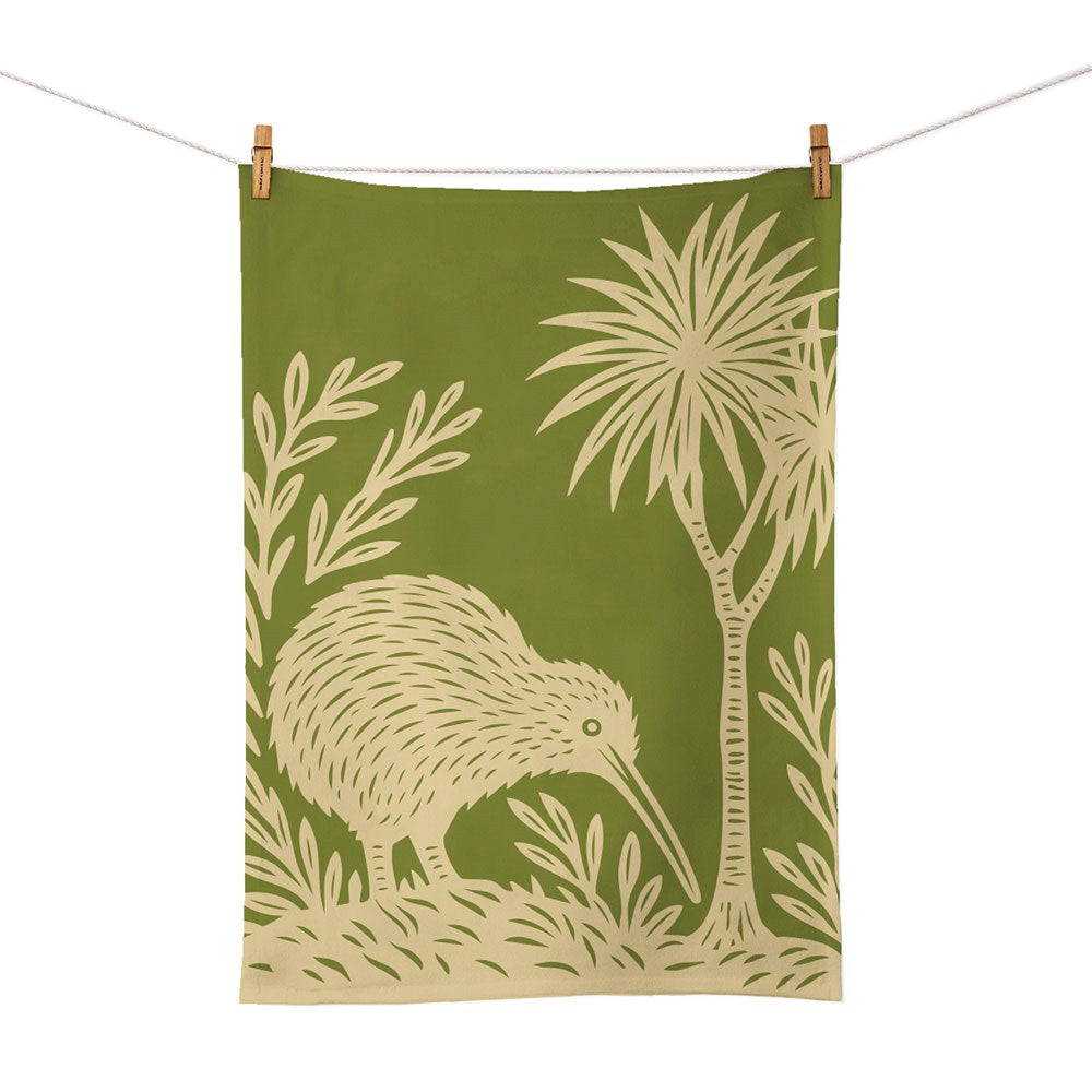 The Woodcut Kiwi Tea Towel is a green cotton towel screen printed with a beige kiwi bird near trees and plants, hanging from a clothesline—celebrating New Zealand’s unique wildlife.