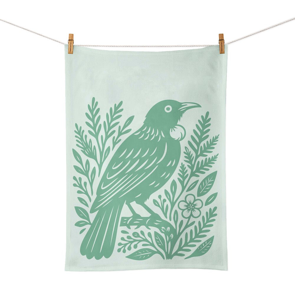 The Woodcut Tui Tea Towel is a light green cotton towel featuring a dark green screen-printed tui bird on a branch, surrounded by leaves and flowers, inspired by New Zealand nature. It is shown hanging from a clothesline with wooden clothespins.