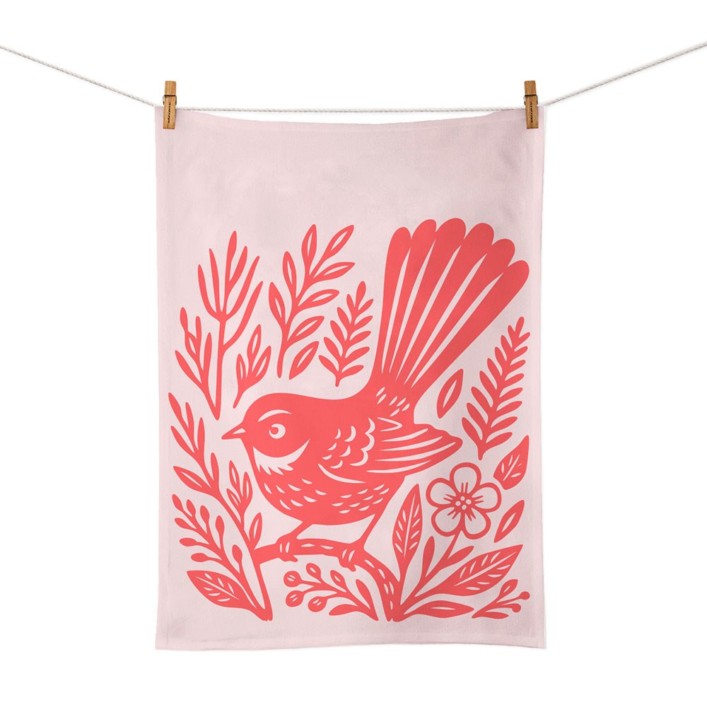 The Woodcut Fantail Tea Towel, made from light-colored cotton and featuring a screen-printed red bird and leaf design inspired by New Zealand, hangs on a clothesline with two wooden clothespins.