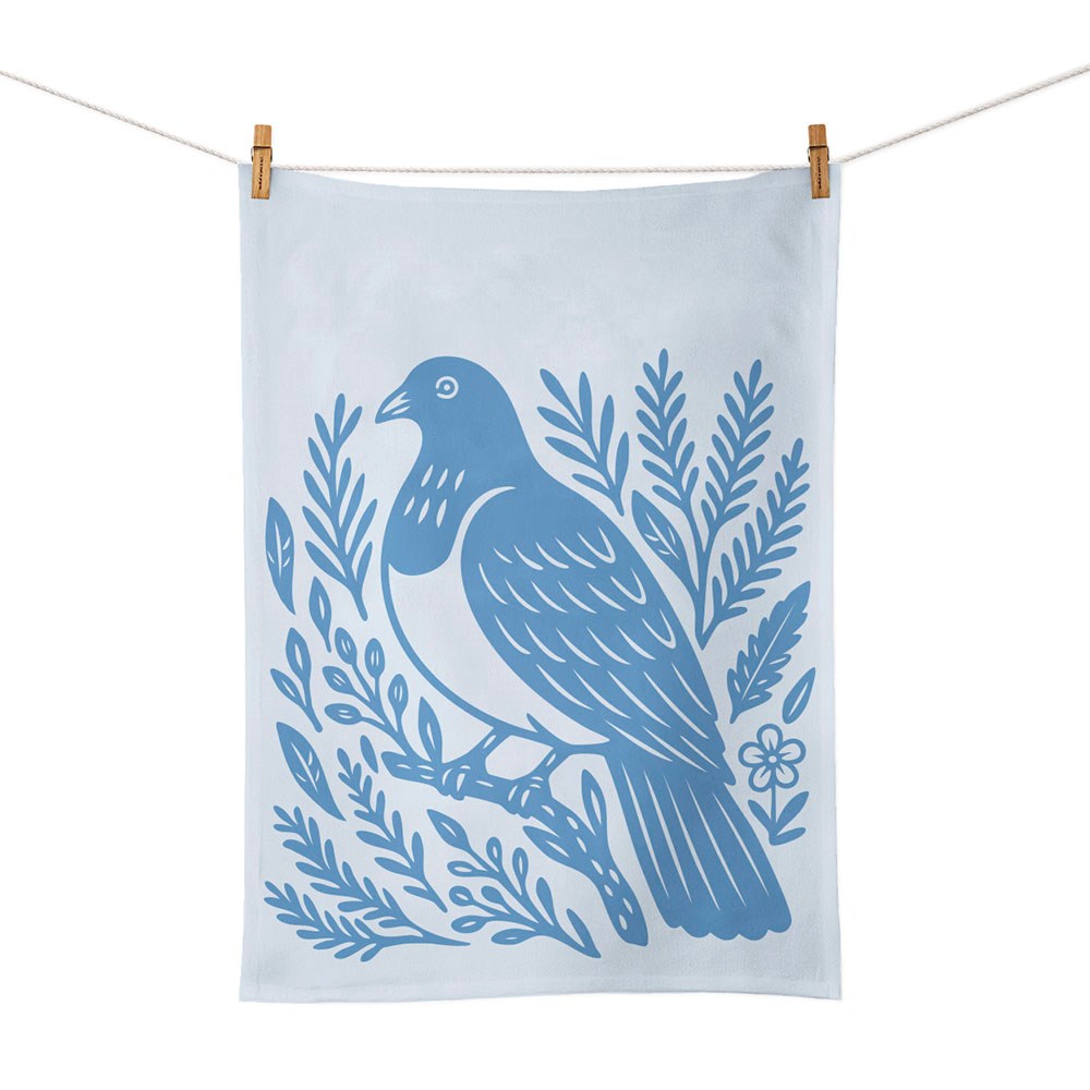 The Woodcut Kereru Tea Towel is a light blue cotton towel with a dark blue bird-and-branch screen print, designed in New Zealand. Shown hanging on a clothesline with two wooden clothespins.