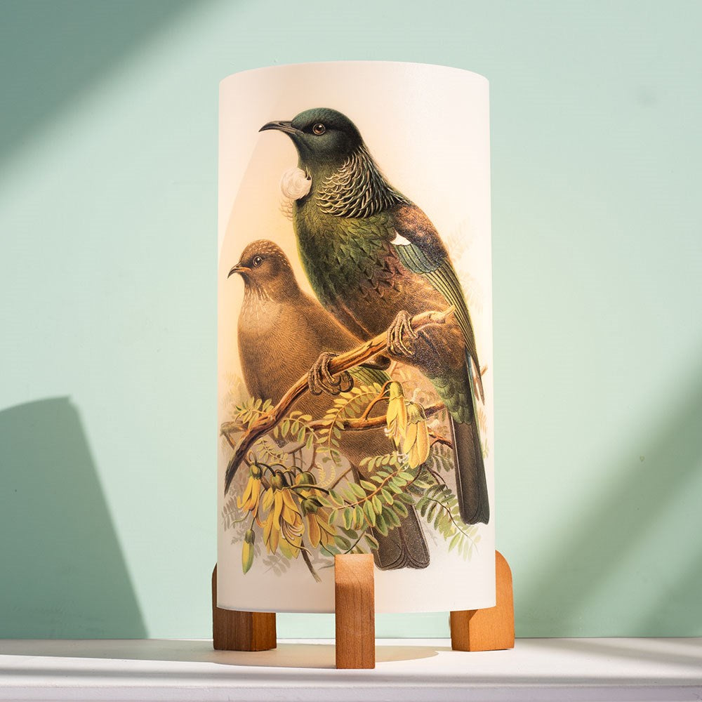 The Buller Tui Table Lamp features a cylindrical shade with two green and brown birds perched on branches, set against a light blue background. Inspired by New Zealand's natural beauty, it sits on wooden feet and is ideal for table lamps.