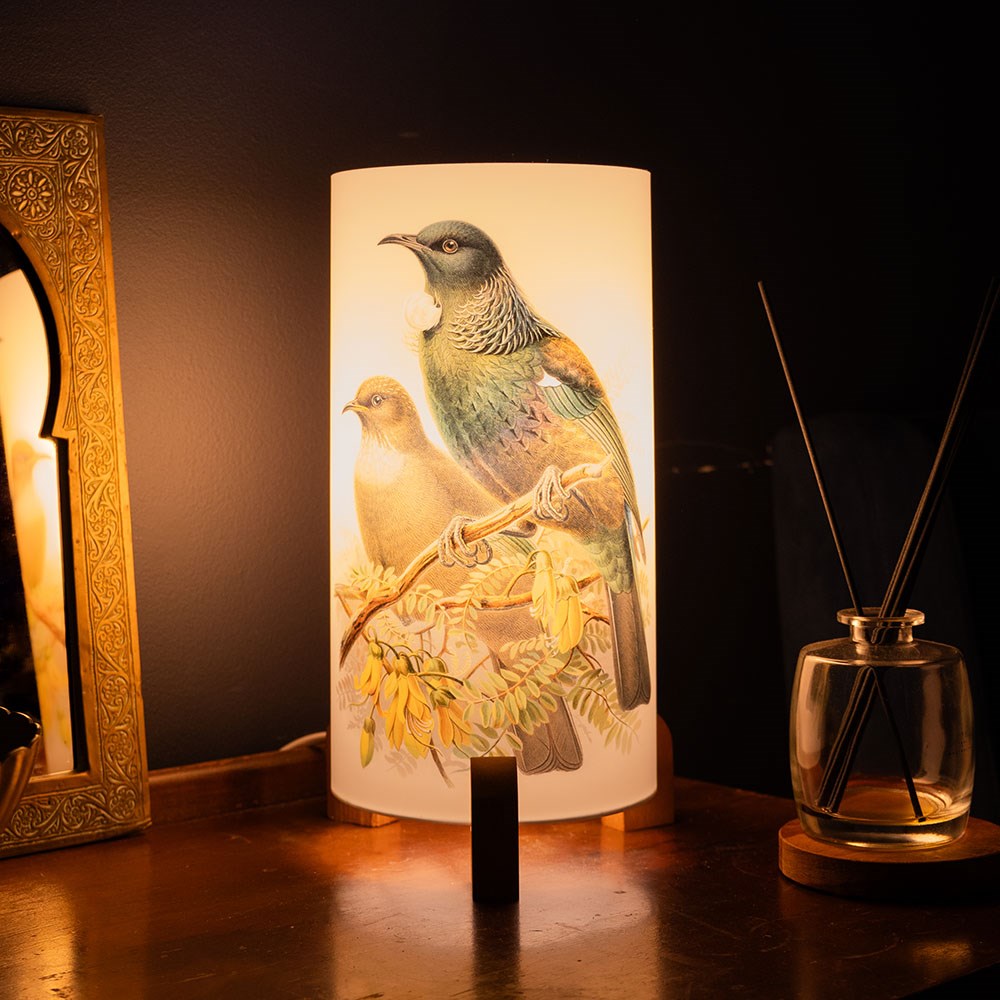 The Buller Tui Table Lamp features wooden feet and an illustrated shade with two birds on branches. It sits on a wooden surface beside a framed mirror and reed diffuser, evoking the mood of New Zealand’s tranquil evenings.