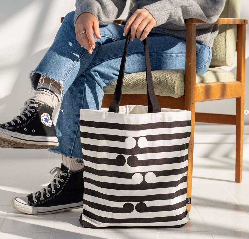 A person in jeans, a gray sweater, and black Converse sits on a chair holding the Gordon Walters - Untitled-No.1 Tote Bag, featuring black horizontal stripes and abstract dot patterns inspired by the artists work.