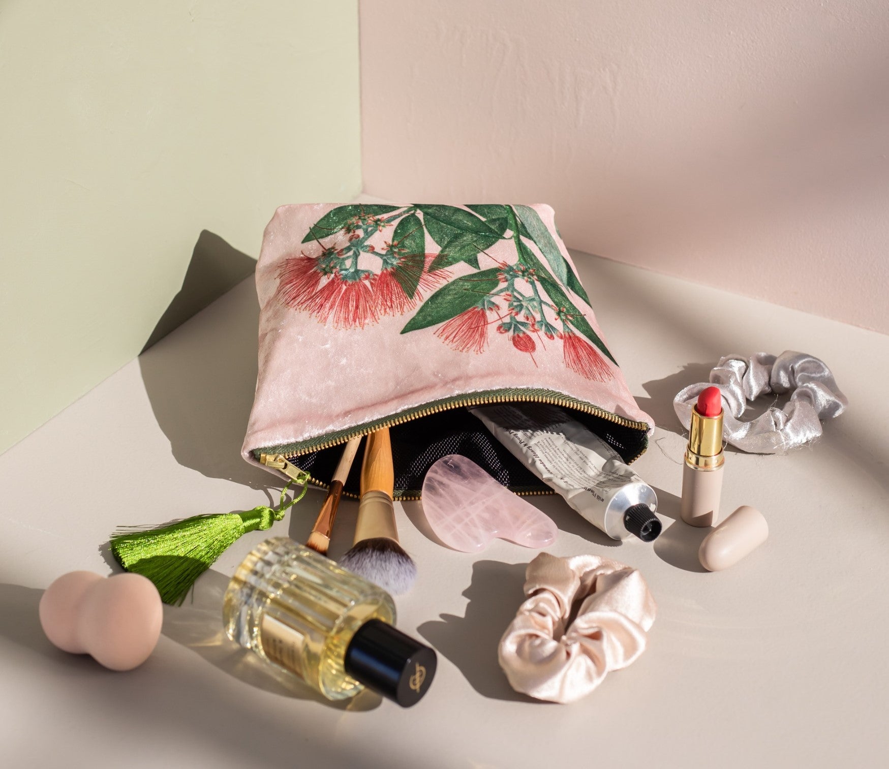 The Vintage Botanical Pohutukawa Velvet Cosmetic Bag, with a pink floral design and green tassel, is open on a light surface among makeup brushes, jade roller, perfume bottle, scrunchie, lipstick, and beauty essentials.
