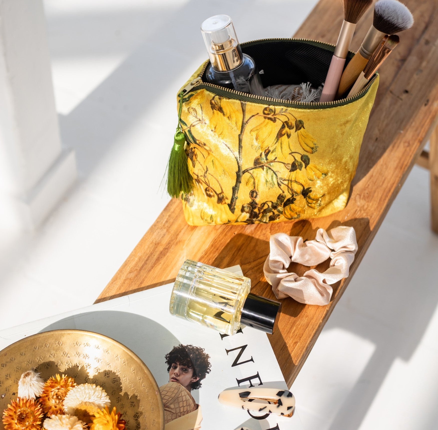 A yellow Vintage Botanical Kowhai Velvet Cosmetic Bag, holding brushes and a bottle, rests on a wooden bench beside a scrunchie, perfume, gold floral dish, hair clip, and magazine. Sunlight creates shadows over this botanical-inspired scene.