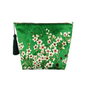 The Manuka Flower Velvet Cosmetic Bag is a green zippered pouch with a dark green tassel, embroidered white and pink Manuka flowers, and green leaves, inspired by New Zealand’s floral beauty.