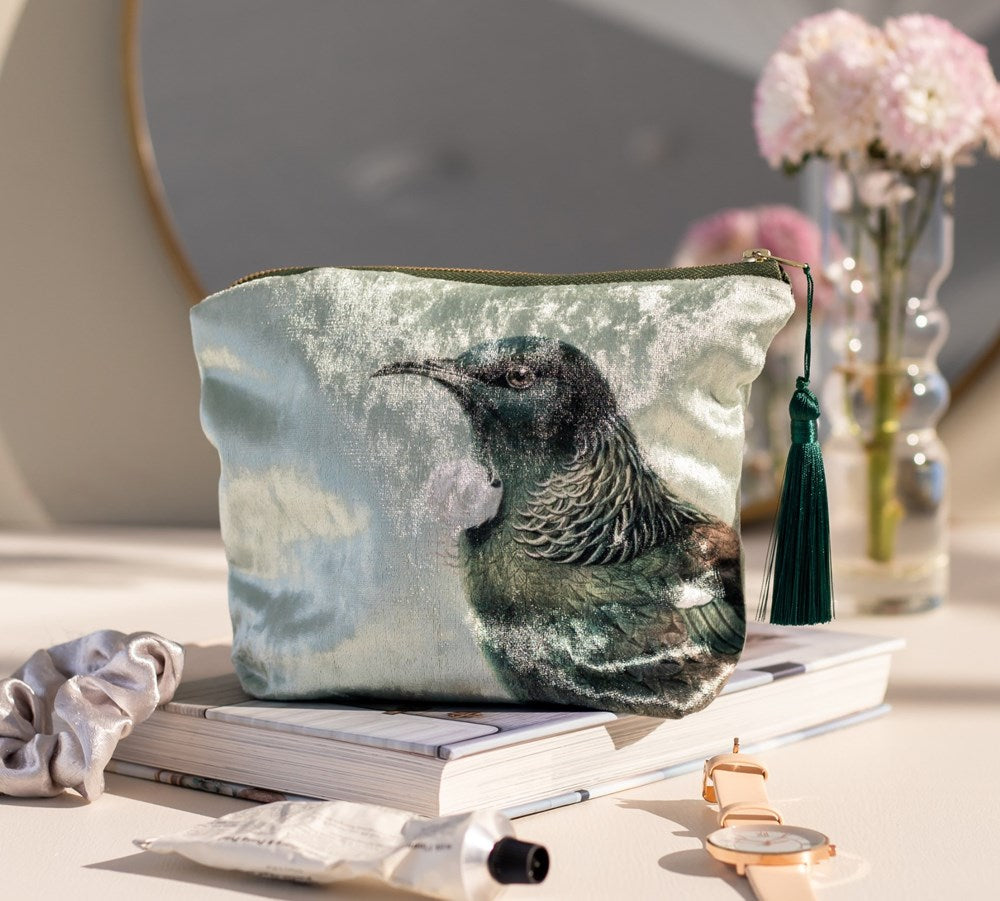 The Hushed Green Tui Velvet Cosmetic Bag, featuring a New Zealand native birds print and green tassel, rests on stacked books beside a scrunchie, watch, and pink flowers—a perfect gift idea illuminated by natural light.