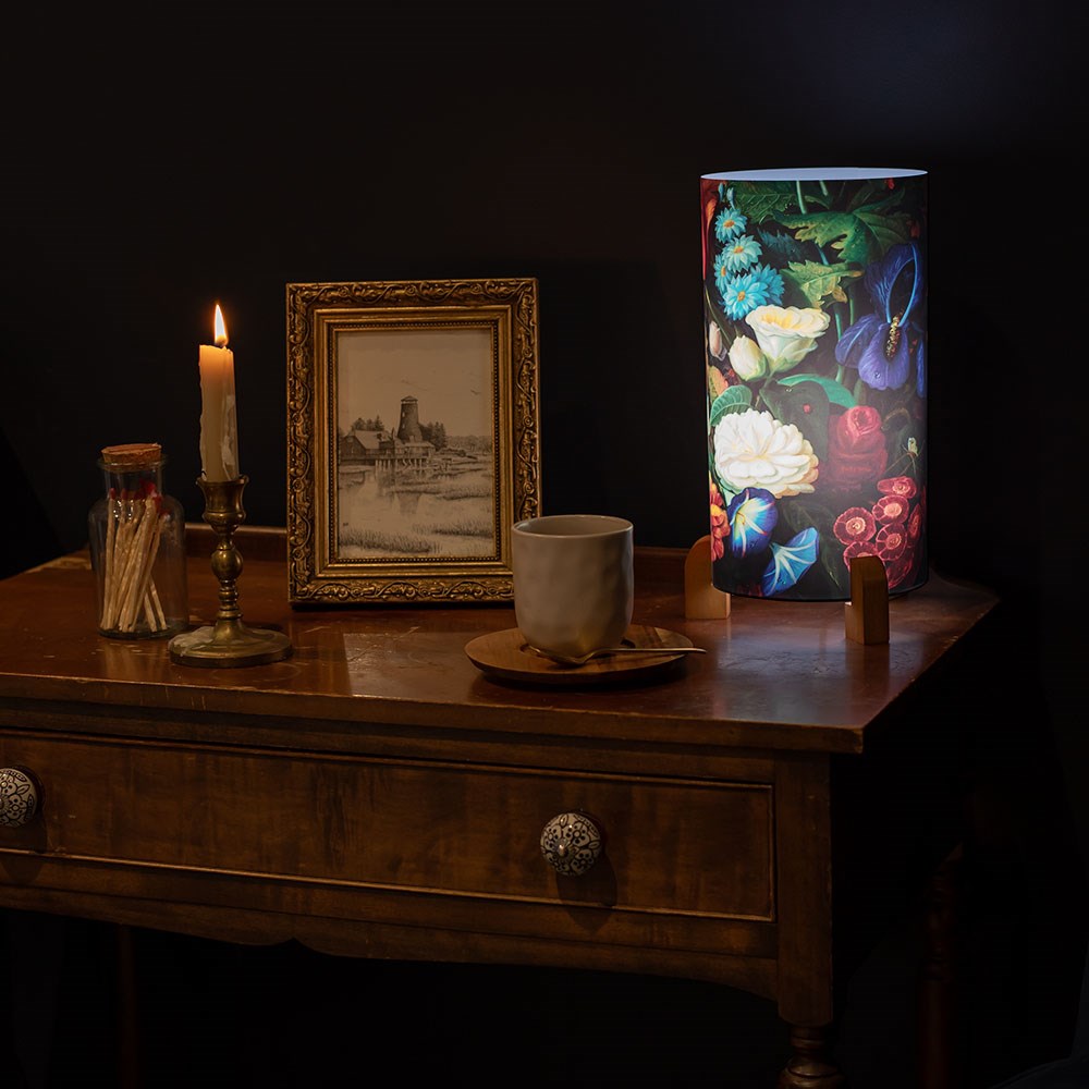 A wooden table with a lit candle, a jar of matches, a framed black-and-white photo, a cup and saucer, and the Vintage Flowers Table Lamp stand out against the dark background—evoking memories distant from vivid images of New Zealand.