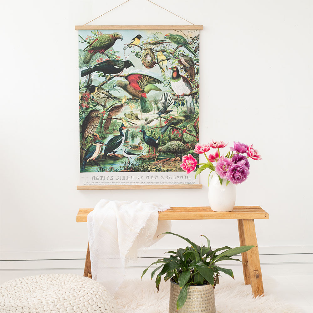 A wooden bench with a white cloth and a vase of pink and purple flowers sits in front of the Native Birds of New Zealand Wall Chart. A green potted plant is on the floor beside the bench.