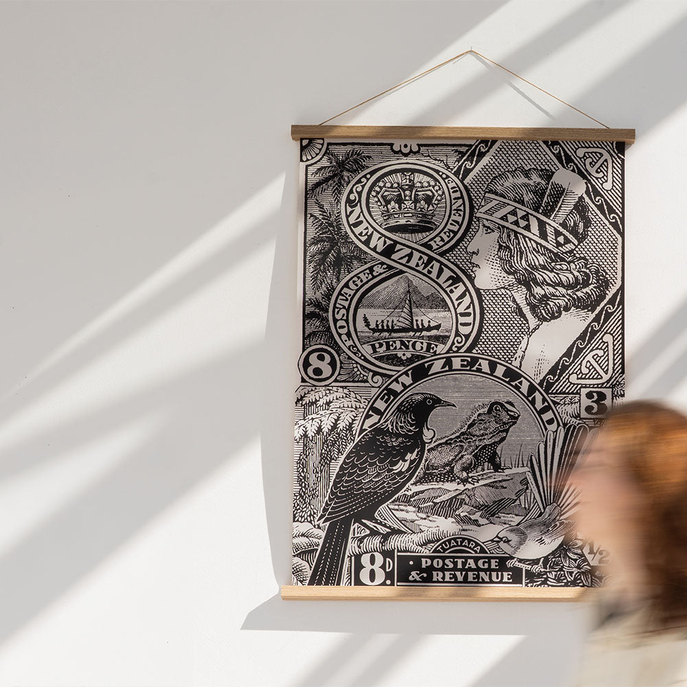 The NZ Stamp Wall Chart features black and white vintage stamp art of a bird, lizard, and portrait. Made in New Zealand, it comes ready to hang. A blurred person walks in front of this striking wall hanging.