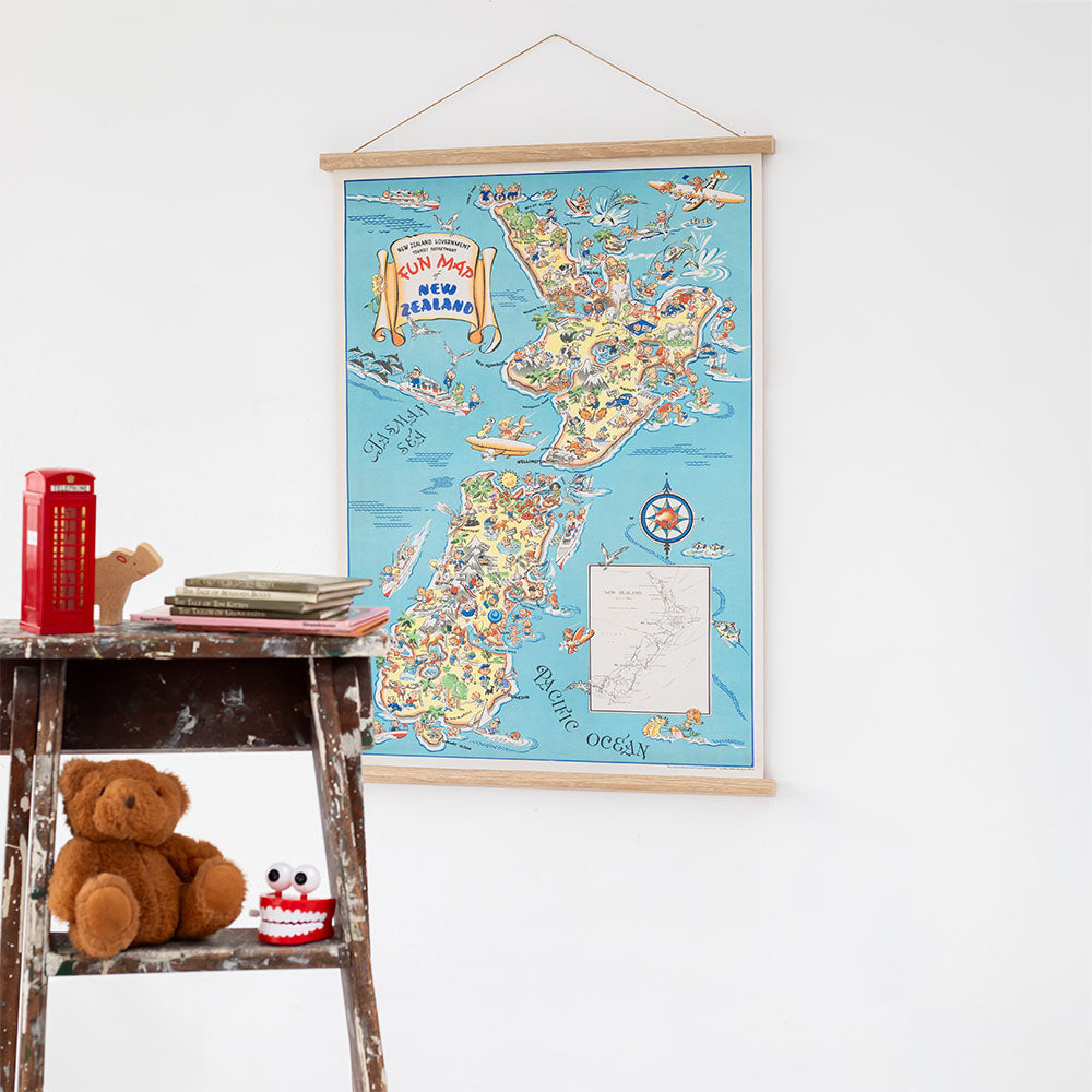 The 1950s Fun Map Wall Chart of New Zealand hangs on a white wall above a small wooden stool holding a brown teddy bear, a red toy bus, a small decorative toy, and a plush toy with large teeth.