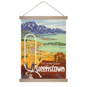 The Queue for Queenstown Wall Chart is a colorful, illustrated New Zealand vintage poster featuring a large Q, the phrase Get in the Queue for Queenstown, scenic mountains, a lake, a town backdrop, and four people in outdoor gear in the foreground.