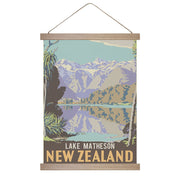 The Lake Matheson Wall Chart is a vintage-style New Zealand poster with illustrated mountains, trees, and lake reflections in cool tones. It hangs by a string with wooden battens and reads Lake Matheson New Zealand at the bottom. Made in NZ.