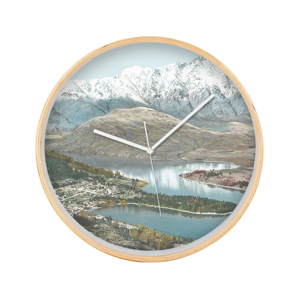 The Queenstown Wooden Frame Clock showcases a vintage print of a lake, small town, and snow-capped mountains. Its white hands move silently, making it ideal for adding calm elegance to any space.