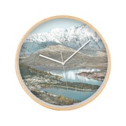 The Queenstown Wooden Frame Clock showcases a vintage print of a lake, small town, and snow-capped mountains. Its white hands move silently, making it ideal for adding calm elegance to any space.