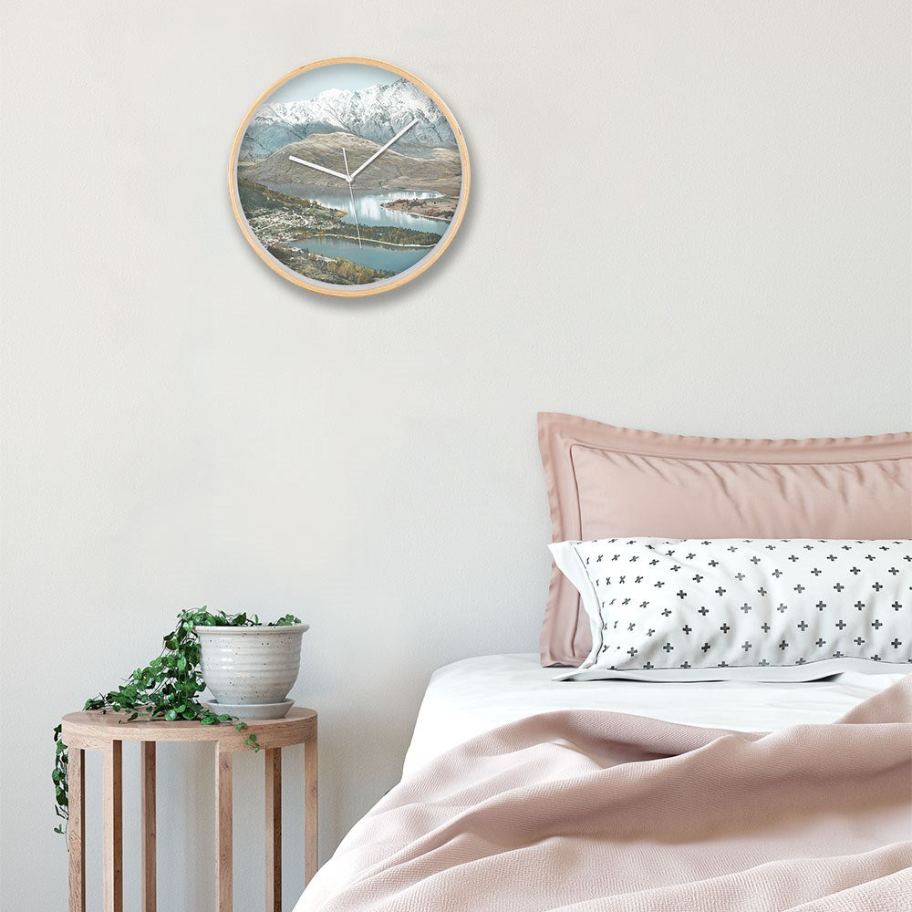 A neatly made bed with light pink bedding sits beside a small wooden side table holding a potted plant. Above the bed, a Queenstown Wooden Frame Clock with silent movement and a mountain lake landscape hangs on the white wall.