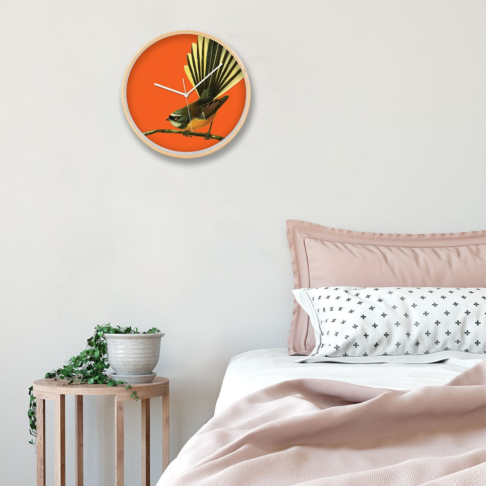 A modern bedroom displays the Bright Fantail Wooden Frame Clock with silent movement above a bed with pink bedding and a cross-patterned pillow, while a small table nearby holds a potted plant.