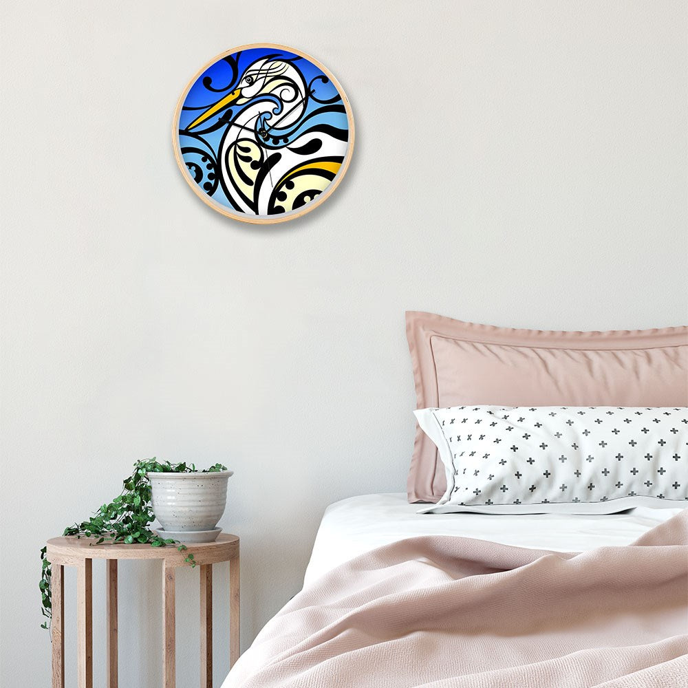 A Shane Hansen - Kotuku Wooden Frame Clock with abstract blue, white, and black bird design hangs above a bed with pink and white bedding. A small wooden side table holds a potted trailing plant.
