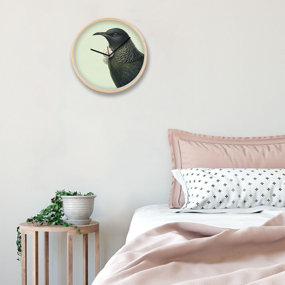 A bedroom corner features a wooden side table with a potted plant, a bed with a pink duvet and patterned pillow, and the Hushed Green Tui Wooden Frame Clock inspired by New Zealand hanging on a light-colored wall.