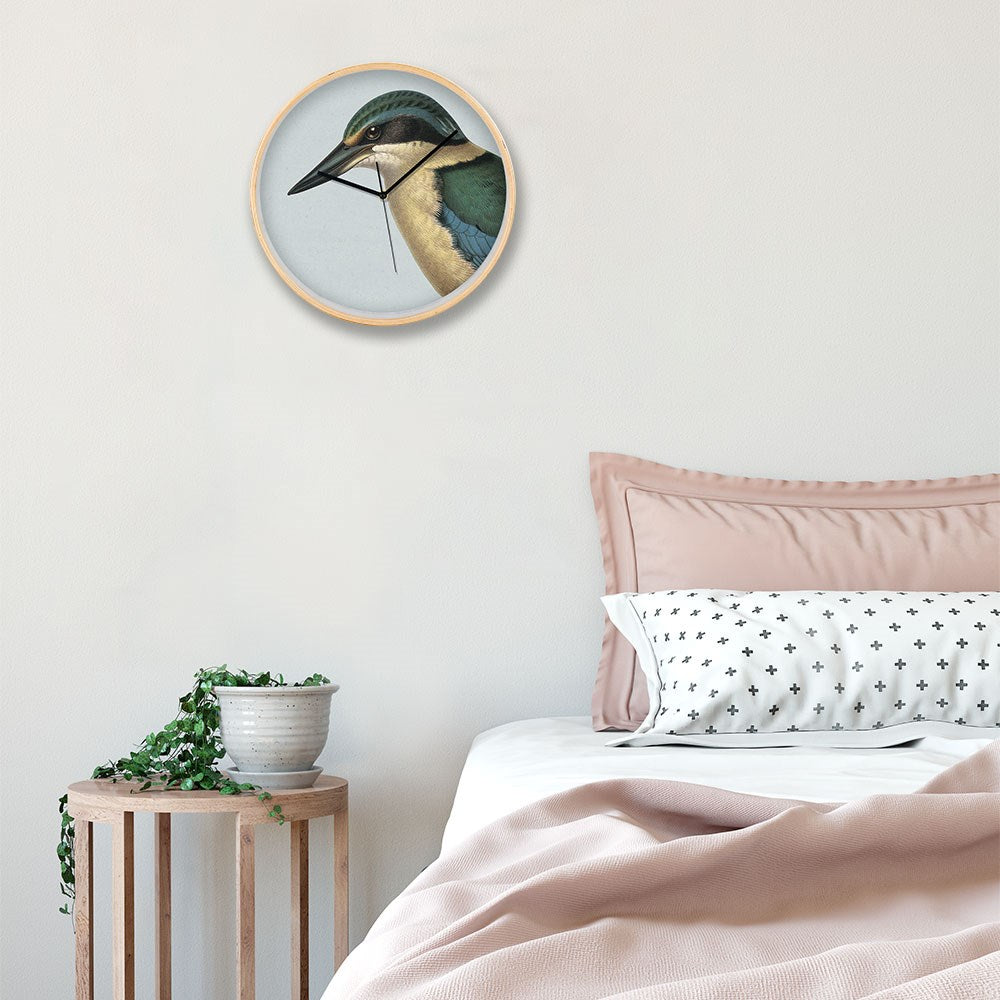 A minimal bedroom features a wooden side table with a potted plant, blush pink bedding, a white pillow with black crosses, and the Hushed Blue Kingfisher Wooden Frame Clock as round wall art.