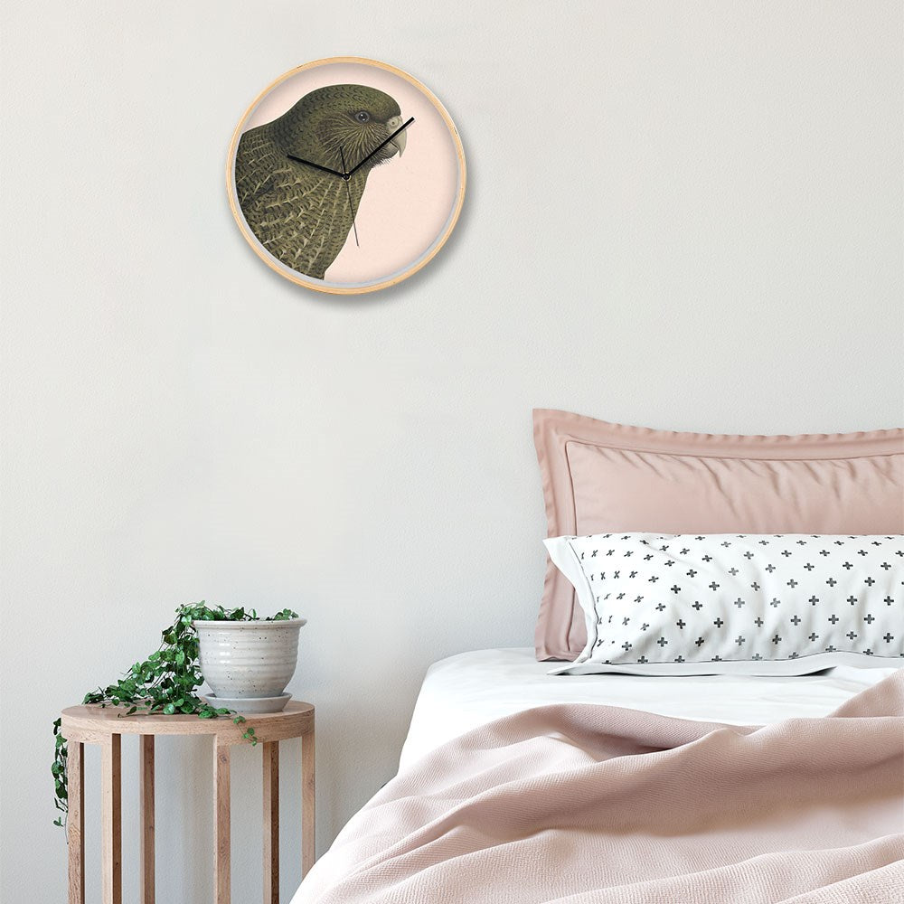 A bedside scene with a wooden stool holding a potted plant, a bed with pink sheets and pillows, and the Hushed Pink Kakapo Wooden Frame Clock featuring New Zealand’s Native Kakapo on a light-colored wall.