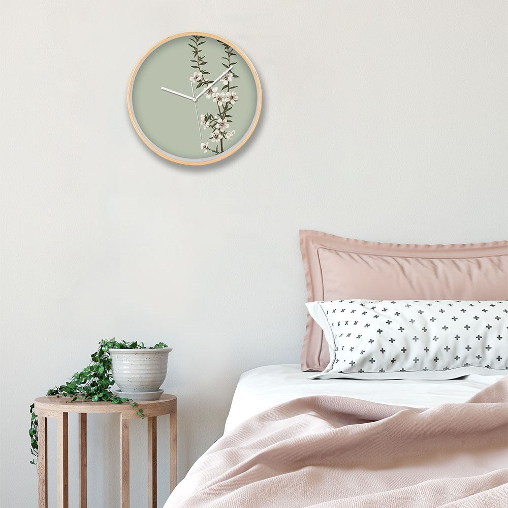 A bedroom corner with a bed, light pink bedding, a white pillow with black crosses, a wooden stool with a potted plant, and the Vintage Botanical Manuka Wooden Frame Clock featuring New Zealand’s native Manuka floral design.
