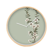 The Vintage Botanical Manuka Wooden Frame Clock features a pale green face, white hands, a silver second hand, and a design of New Zealand’s native Manuka branch with white flowers and green leaves on the right side.