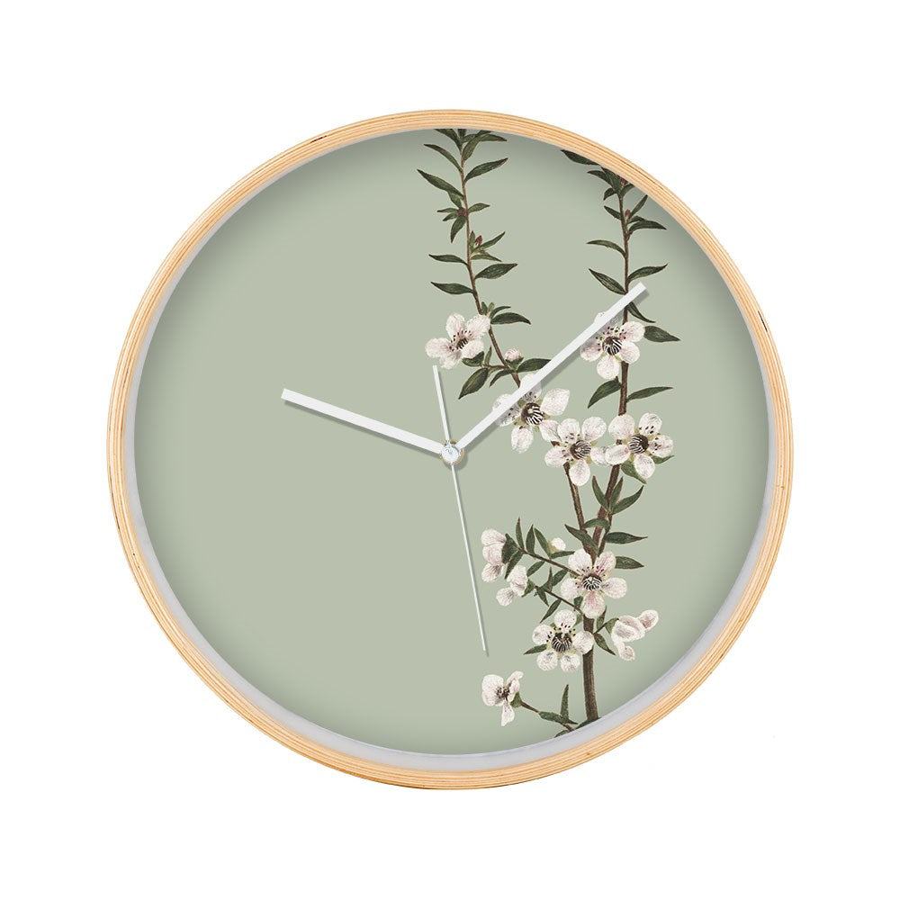 The Vintage Botanical Manuka Wooden Frame Clock features a pale green face, white hands, a silver second hand, and a design of New Zealand’s native Manuka branch with white flowers and green leaves on the right side.
