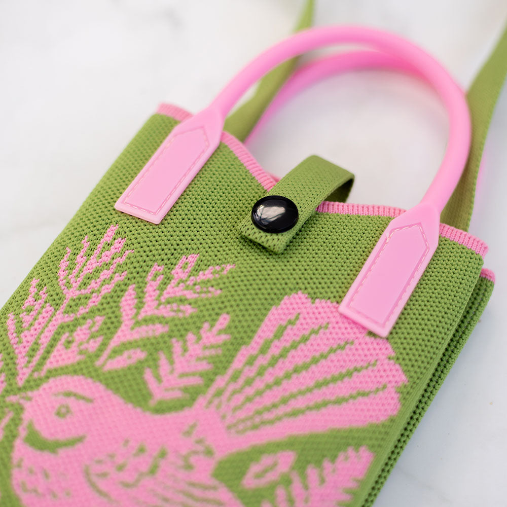 The Woodcut Fantail Cross-body Mini Bag is a green fabric mini bag featuring pink handles and a pink native bird artwork on the front. This lightweight cross-body bag has a black button closure and is shown in the photo on a white surface.