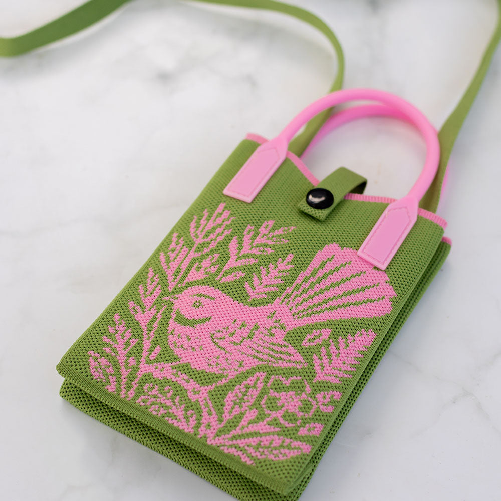 The Woodcut Fantail Cross-body Mini Bag is a small green fabric handbag featuring pink handles and a pink cross-stitch pattern of native bird artwork with leaves. This lightweight bag has a button closure and is displayed on a white marble surface.