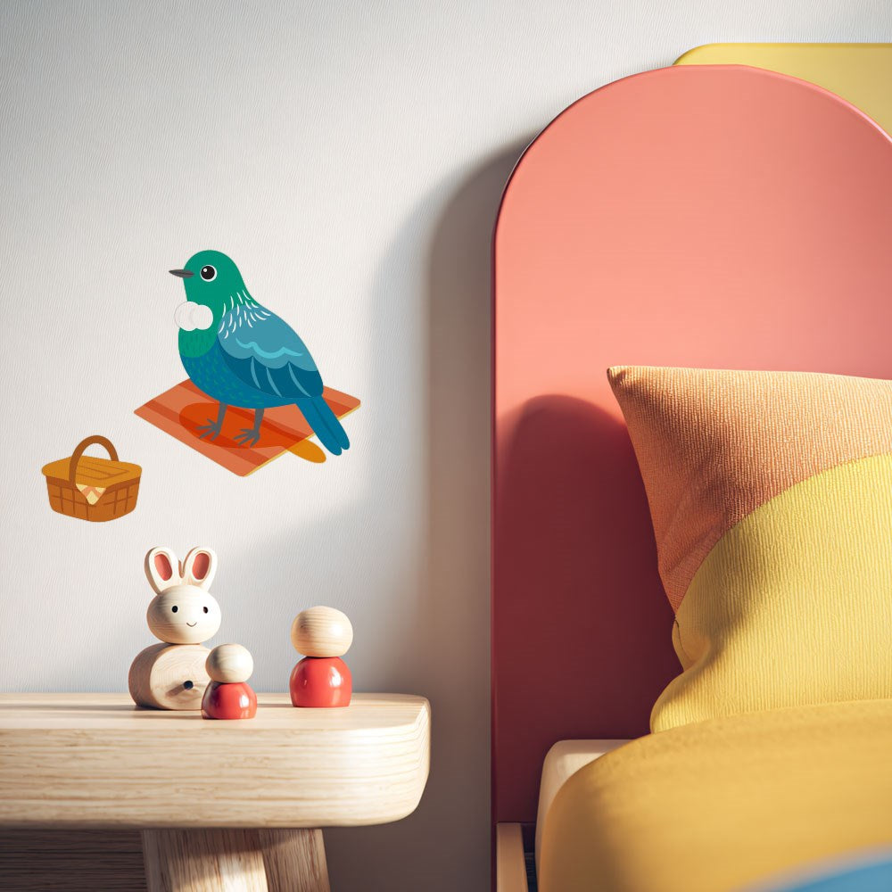 A children’s bedroom scene features a wooden nightstand with toy figures, a white rabbit, and the Tui Wall Decal—showing a bird, picnic basket, and orange plaid blanket—on the wall. The bed has a colorful pillow and matte fabric finish.