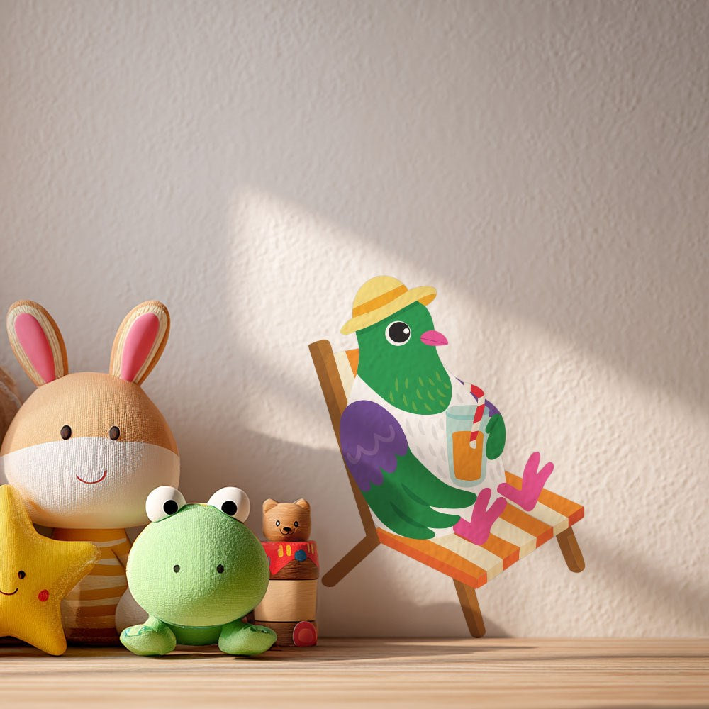 A plush bunny, yellow star, green frog, and small cat figurine sit on a wooden surface. On the wall is the Kereru Wall Decal—featuring a pigeon in a hat sipping from a cup—just peel and stick for easy decor.