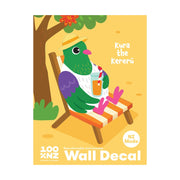 An illustrated green bird with a hat and sunglasses relaxes on a striped deck chair under a tree, holding a drink. Text: “Kereru Wall Decal,” “NZ Made,” “Wall Decal.” Peel & stick, matte fabric finish. Yellow background.