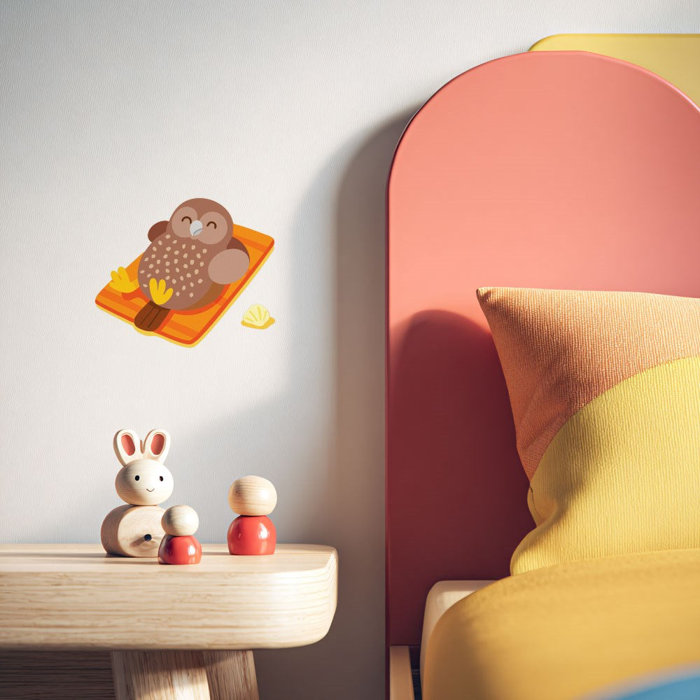 A kids’ bedroom with a wooden nightstand, bunny and figure toys, and the Ruru Wall Decal—an owl on a branch in matte fabric—on the wall. The bed features a yellow-orange pillow and pink headboard.