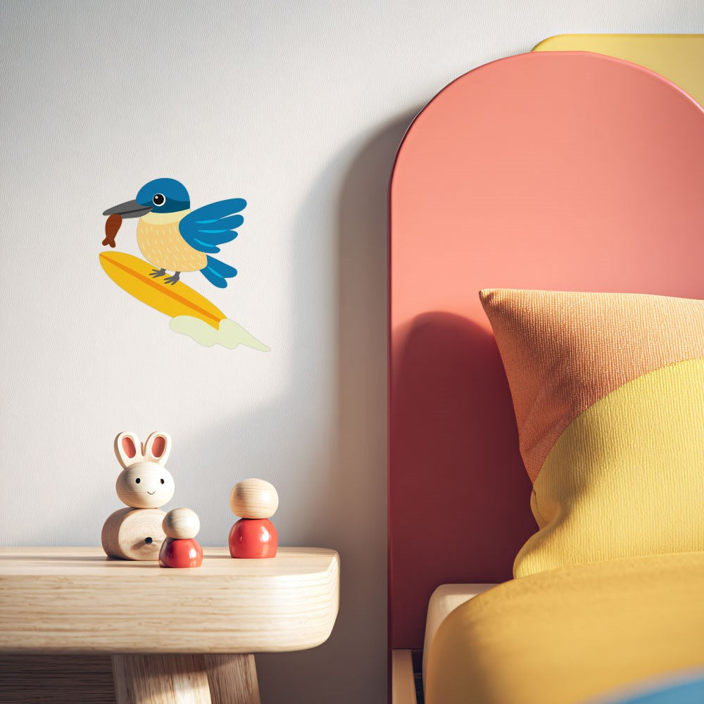 A children’s bedroom features a wooden bedside table with two wooden toys, a bed with colorful bedding, and the Kingfisher Wall Decal—a peel and stick matte fabric wall art of a blue bird flying above a yellow surfboard shape.